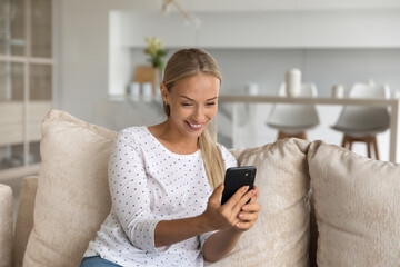 Happy excited millennial woman having nice video call talk. Mobile phone user sitting on couch at home, looking at smartphone screen, laughing, chatting online, taking selfie, getting good news