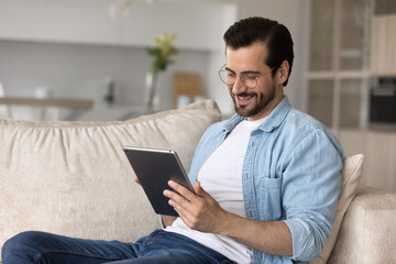Happy user holding digital device, resting on couch, relaxing at home. Relaxed millennial man using...