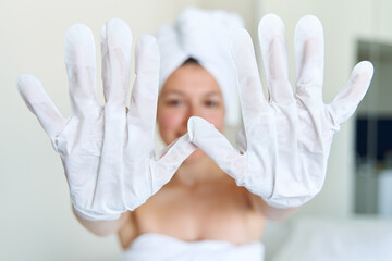 After a shower, a girl wrapped in a towel uses cosmetic gloves to moisturize the skin of her hands. Cosmetic trends for body care at home