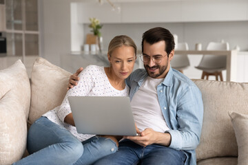 Happy millennial couple focused on laptop sitting together on sofa at home, hugging, using app on...