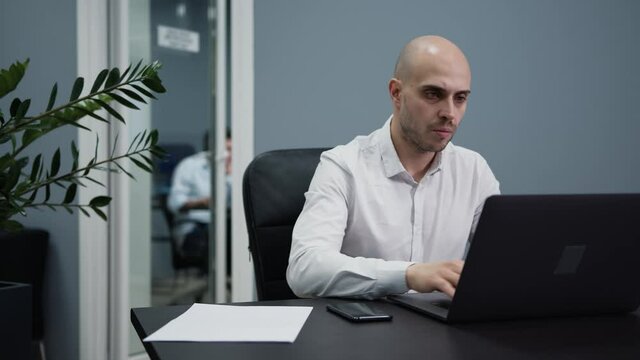 Businessman with laptop, checking phone on the table, busy working with laptop. Young manager in moden office with colleagues on background, devices and papers on the table