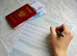 a woman's hand holds a pen and begins to fill out an auto insurance form, next to it lies a Russian passport and money
