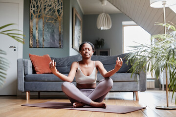 Full length portrait of young African-American woman meditating at home while sitting in lotus position with eyes closed