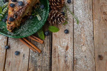 Piece of tasty cake with blueberries on wooden table with cones, cinnamon and mint. New Year Eve vintage meal