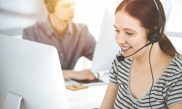 Casual dressed young woman using headset and computer while talking with customers online. Group of operators at work in sunny office. Call center