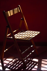 A wooden chair stands in front of a red wall and the shadow of the chair falls on the floor, in a...