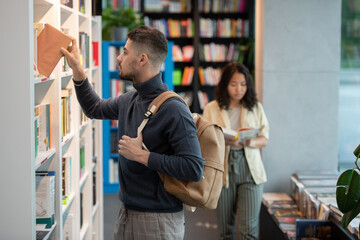 Adolescent man in casualwear taking book from bookshelf in college library