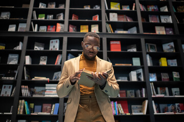 Young serious man in smart casualwear and eyeglasses reading book in front of camera against...