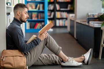 Serious young student in casualwear sitting against bookshelves and reading one of books