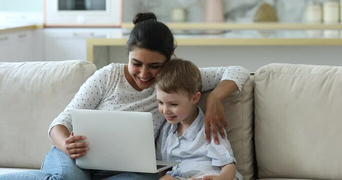 Indian nanny and little boy sit on sofa at home watch cartoons on laptop enjoy educational online program for preschool education spend free time on internet. Modern tech, parental control concept