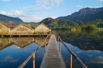 Fototapeta na wymiar A long pier leading to the wooden boat houses on lake Kochelsee with the scenic Bavarian Alps against the blue sky reflected in the lake in the background (Kochel am See, Bavaria, Germany)