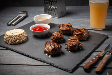 Grilled pork medallions with cheesy salad, ketchup and mustard on dark stone board. Served with glass of beer.