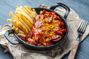 Currywurst with french fries in black pan on craft paper on wooden table.