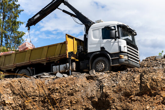 Side bottom pov big modern truck with mounted self loading crane arm boom unloading pallet of new bricks at countryside rural forest suburban construction site. Building materials delivery service