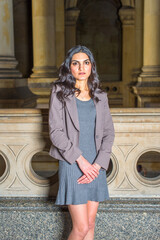 Dressing in a gray knit dress and a jacket, a young businesswoman is standing in an office hallway, confidently looking at you.