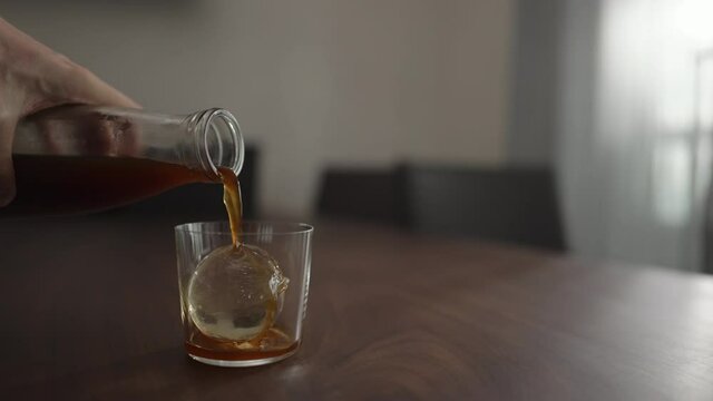 Slow motion pour cold brew coffee over ice ball in tumbler glass on walnut table