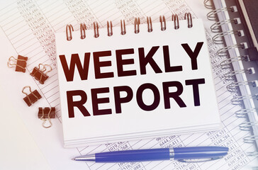 On the table is a pen and a notebook with inscriptions - WEEKLY REPORT