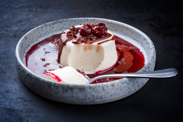Modern style traditional blancmange almond pudding with cherry, chocolate cream and sour cherry gelee compote coulis served as close-up in a Nordic design plate on a black board