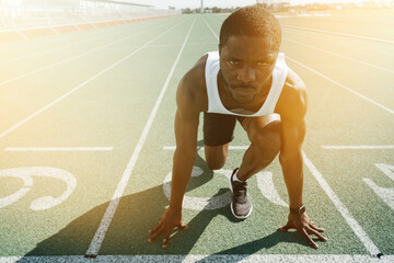 A dark-skinned male track and field athlete with a serious face stands on the start line leaning on...