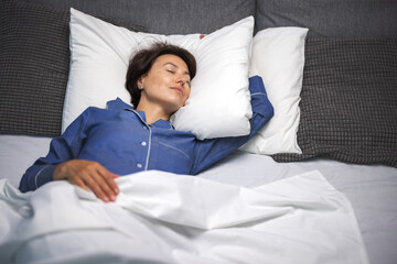 Mature woman in pajamas enjoying sweet dreams on comfy bed with soft white linen. Peaceful sleep of...