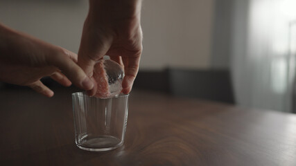 man put ice ball into tumbler gllass on walnut table