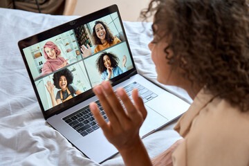 Fototapeta na wymiar Young African American teen girl lying on bed having virtual meeting waving hand chatting with friends during online video call at home having fun using laptop computer. Over shoulder view