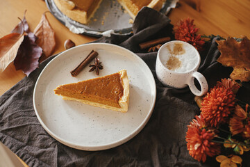 Happy Thanksgiving. Pumpkin pie slice on modern plate and hot coffee on rustic table with linen napkin, autumn flowers and leaves, anise and cinnamon. Homemade pumpkin tart recipe.