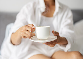 Close up of white ceramic cup with hot aroma coffee in female hands. Mature lady in sleepwear drinking morning beverage while sitting on bed.