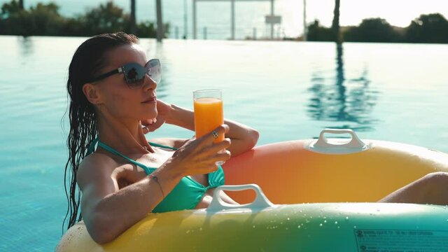 In swimming pool serene 30s brunette woman in bikini and sunglasses sits inside rubber circle hold glass, drinks cocktail or orange juice, enjoy summer holidays, leisure, vacation pleasure concept