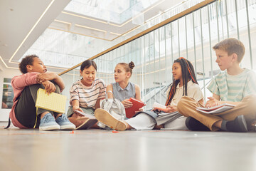 Full length portrait of diverse group of children sitting on floor in school and chatting while waiting in hall