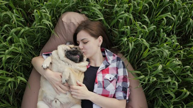 Woman lies with a pug in the field on bag chair, kiss and hugs dog, playing in nature, top view