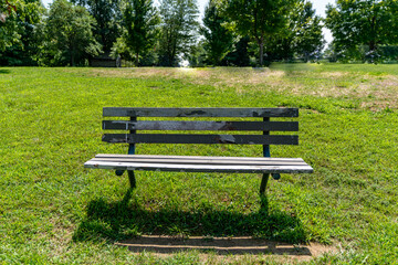 An old deteriorating wood and metal park bench in a grass field on a sunny summer day surrounded by tall trees.