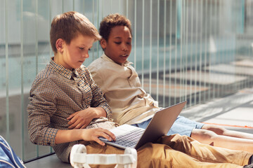 Portrait of two schoolboys using laptop while sitting on floor in school lit by sunlight, copy space