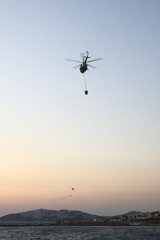 A helicopter of the fire service with a fire fighting bucket is taking part in putting out a fire. A helicopter with a red basket is lowered over the sea to catch water.
