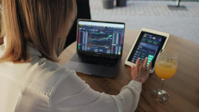 Woman trader trades and analyzes the cryptocurrency market, invests money while sitting in a cafe, drinking a cool cocktail, use laptop and tablet