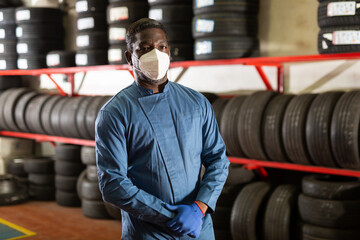 Fototapeta na wymiar Portrait of professional afro american auto mechanic in face mask posing near car tires at auto service