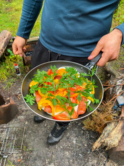 Breakfast or lunch outdoors. A man cooked fried eggs in a frying pan outdoors. Eggs, salt, sausages, peppers, tomatoes, parsley and herbs. Very tasty and healthy. Lots of protein