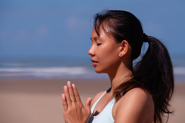 people, fitness, sport and healthy lifestyle concept - close up portrait of young asian woman making meditation on tropical beach with blue sky background