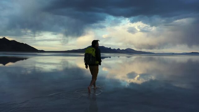 Silhouette of Man With Backpack Walking in Surreal Landscape of Salt Flats With Stormy Clouds and Sky Mirror Reflection on Water, Cinematic Approaching View