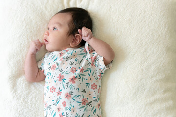 Top view of cute little Asian baby girl, wearing a pretty floral dress, lying on white bed and looking out