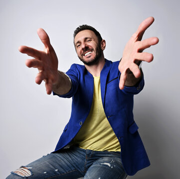 Portrait Of Friendly Smiling Adult Man In Casual Clothing Sitting On Stool Holding Out His Hands, Fingers Spread Out, Going To Hug Or Take Something Over White Background