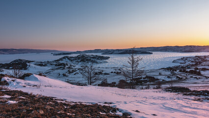 Dawn over a frozen lake. The sky over the mountain range is highlighted in orange. Sun glare on the ice. Houses of the tourist base on the shore. Snow on the ground and bare trees. Baikal 