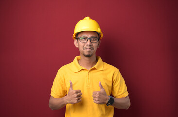 Handsome Asian worker man wearing helmet smiling and thumbs up over isolated red background.