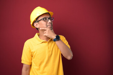 Handsome Asian worker man wearing helmet thinking over with hand on chin against yellow background.