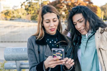 Two beautiful Latin women sharing cell phone messages while laughing in a public park. Technology and communications concept.