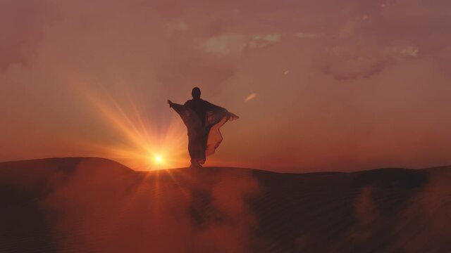 Young woman in traditional Abaya dress and in hijab enjoys nature in the desert at sunset. Woman rising her hands stand at the peak of deserts mountains with fog and wind