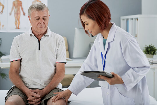 Physiotherapist with tablet computer in hands touching knee of senior patient who had bad injury