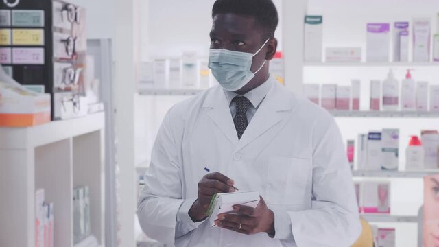 Portrait of African-american male pharmacist in face mask who standing in salesroom of drugstore 