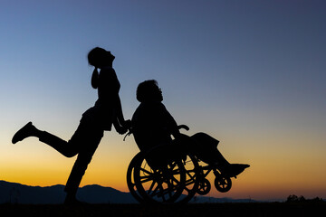 Disabled handicapped young man in wheelchair with his care helper walking in sunset.Silhouette