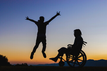 Disabled handicapped young man in wheelchair with his care helper jumping in sunset.Silhouette